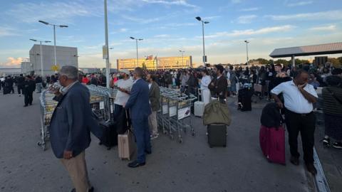Large crowd of passengers with luggage and trolleys gathered outside an airport terminal building under evening light, some standing and waiting while others sit on baggage carts.