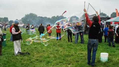 People at Strawberry Fair. One is holding a large bubble set while others observe in front of foot trucks on a grassy field. It is cloudy.