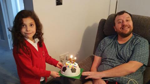 A young girl in a red jumper presents a man sitting in a chair with a football birthday cake with candles in the shape of a 44. 