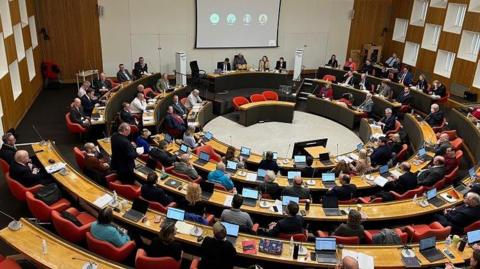 The council in session. There are five elevated tiers of circles of wooden desks and red chairs which councillors are sitting in. There is a single row of four councillors on ground level and there is a visual screen behind them.