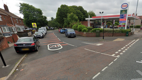 The junction of Meadowside - a residential street - and the A690 in Sunderland. There are several cars parked half on the pavement on both sides. On the corner is a petrol station with a large Esso sign.