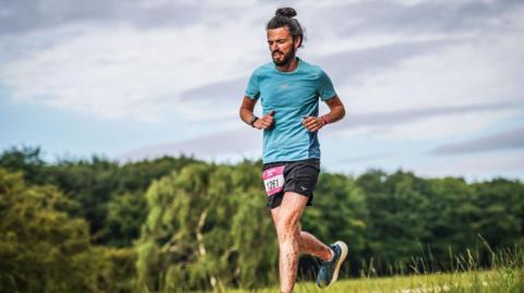 A runner moving captured mid‑stride on a grassy path. He is wearing a turquoise short‑sleeved top, black running shorts and running shoes and has a race number bib is attached to the front of his shorts.
