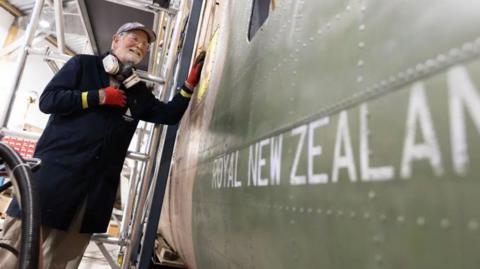 A volunteer standing beside the aircraft, resting one hand on its side. The older man is wearing a long navy work coat, red gloves and a breathing ventilator. On the side of the plane it reads 'Royal New Zealand' before the words are cropped out.