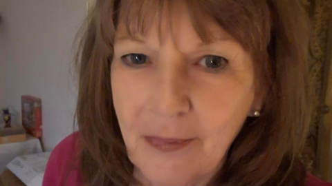 A woman with shoulder-length, light brown hair taking a selfie. She is wearing a red top and is stood in a white room