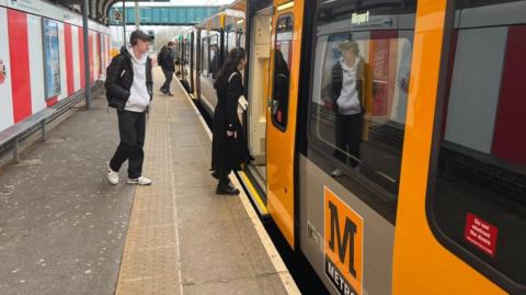 Two men and a woman board a black and yellow Metro train towards Newcastle Airport from Platform 2 at the Stadium of Light station in Sunderland. 