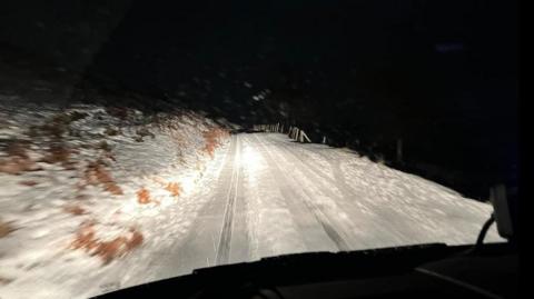 View out of a car windscreen onto a snowy road. The road is covered with snow with two tracks visible, and a snow-covered bank can be seen to the left hand side, to the right all is darkness.