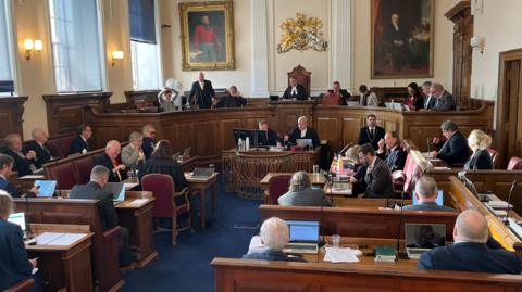 A number of people sitting in suits on wooden benches facing forward, with a top bench with people sitting looking down, in a court setting. 