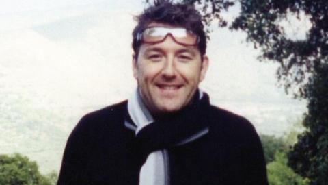 Adam Pickles is standing outside, with trees and mountains behind him. He is wearing a dark coloured top and a white scarf and he is smiling, with glasses worn on his head.