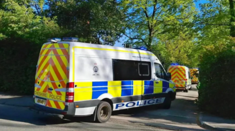 A row of white police vans with blue, yellow and red markings entering a gap in a high hedge