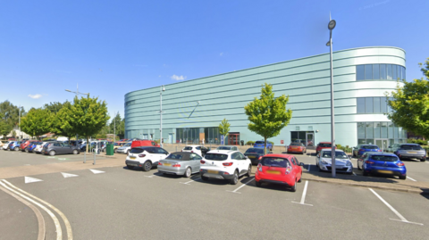There are cars parked in front of a long rectangular green-coloured building with curved ends. There are trees in the distance.
