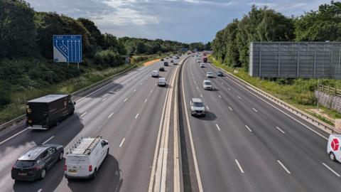 A an aerial shot of a motorway, with traffic on both carriageways. The surface of the road looks new and in good condition. 