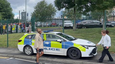 A police car parked outside a school as people walk past.