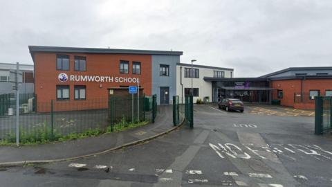 View from the road of Rumworth School. It is a modern red-brick building with grey and purple signage. Reception is visible from afar. The car park gates are open and there is one parked car. 