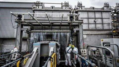 The Magnox Swarf Storage Silo, a futuristic looking factory type building at Sellafield. Two people in yellow helmets and hazard protection suits are working with their backs to the camera. The building is metal and concrete with no windows or obvious openings. Pipe work and industrial equipment can be seen all over the image.