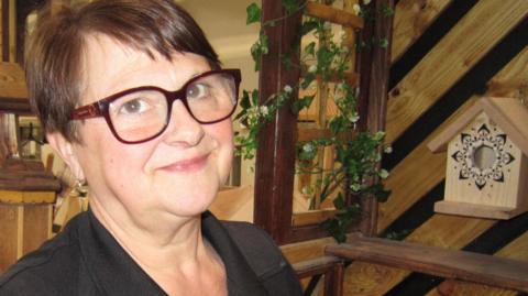 Nichola Joynes, standing in a prison workshop, wearing a black T-shirt, with writing on. She has on glasses and a lanyard round her neck. She has short dark hair. A bird box is to the right and wooden items behind her. 