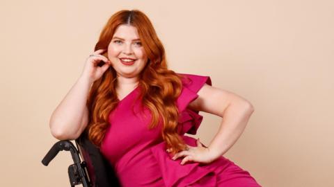 Jenni Pettican posing for a picture in her wheelchair. She has turned on to her right side and is wearing a bright pink coloured dress. She has long red hair and is looking directly at the camera and smiling.
