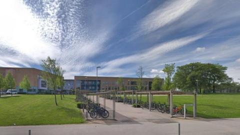 A three-storey building with dozens of stationary bikes parked in sheds. There are trees surrounding the building and the sky is blue. 