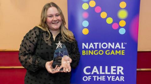 A woman with long blonde hair wearing a black dress poses at the camera and smiles while holding a trophy. She stands next to a board that says, 'National Bingo Game Caller of the Year'. She 