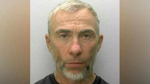 A police mugshot of James Holder. He has cropped grey hair and a short grey beard and is looking directly ahead.
