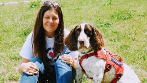 Kerry Snuggs sits in a grass field with her liver and white English springer spaniel. She is looking at the camera, smiling, and has long dark brown hair. She has a pair of large tortoiseshell sunglasses on top of her head and wears a white short sleeved cotton t shirt which has a red heart logo on the front and a pair of blue denim jeans. Her dog sits by her side, also looking at the camera and wears a red harness, which has a badge on it reading 'PTSD Dog'