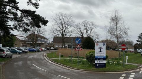 General view of Cottenham Village College from the road which leads up to the school and has parked vehicles outside. There is a sign at the end of the road which has the school name and tree logo on, and says 'welcome'.