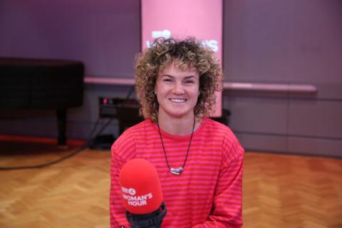  Ellie Kildunne smiles at the camera. She is wearing a red striped, long-sleeve top. She sits in-front of a red BBC Woman's Hour microphone.