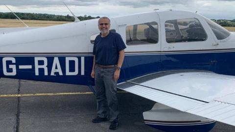A man with a beard and sunglasses is seen standing in front of a blue and white plane