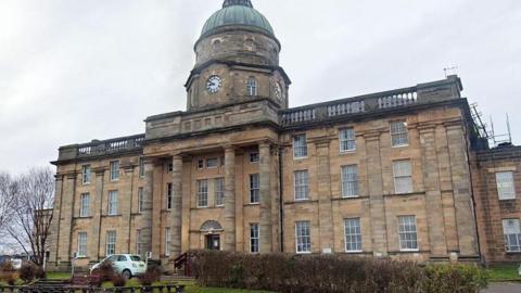 An exterior shot of Dr Gray's on a grey cloudy day, the sandstone building with four columns rises up to a copper dome above an old clock face.