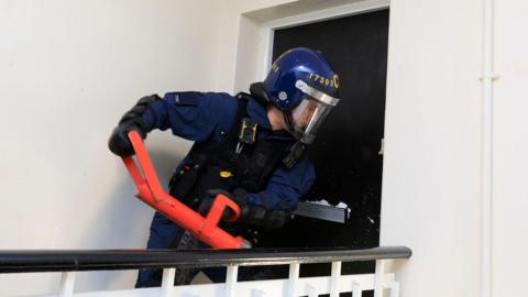 Police officer wearing riot gear force open a front door with a large metal instrument during a raid.