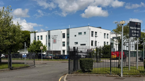 Photograph of St Cuthbert's RC High School in Rochdale. The image shows the school gates, with the large white-clad building in the background.
