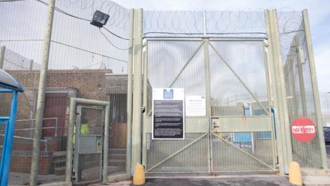 A prison gate with barbed wire and fencing running alongside. Various signs saying 'no entry' and 'HMP Erlestoke' also evident.