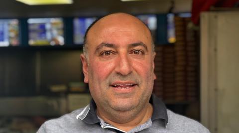 A head and shoulders image of Nuri Ozmicco. He is wearing a grey polo shirt and smiling while looking into the camera.