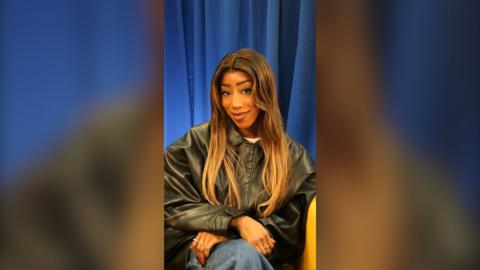 A woman with brown hair and a black jacket sits in a chair.