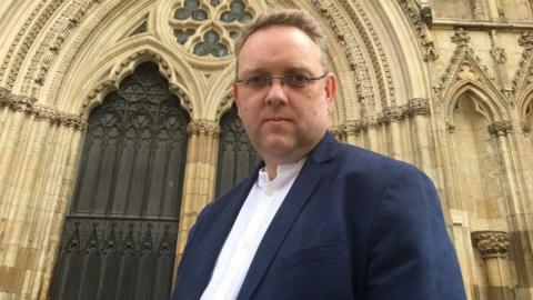 A middle-aged man with glasses, a white shirt and navy suit jacket looks into the camera as he stands against the backdrop of a huge church, which could be a cathedral.