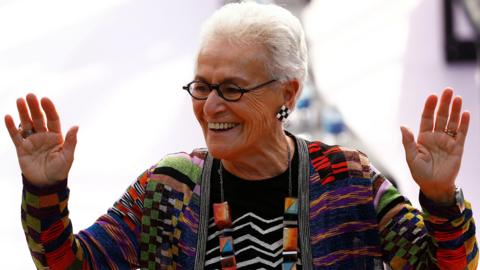 Rosita Missoni, wearing a Missoni cardigan in a distinctive rainbow pattern with a bold statement necklace and earrings, waves at viewers during a dashion show in 2017