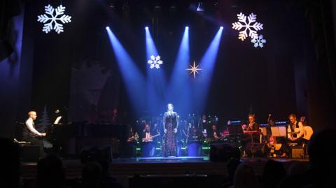 A woman in a sparkly dress stands under a spotlight on a dark stage, she is is surrounded by musicians.