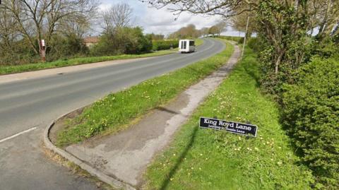 The junction of King Royd Lane and Doncaster Road in Ackworth, West Yorkshire in spring. A white van is in the distance, on a grey road with green grass verges. There are trees with bare branches on both sides of the road and a bus stop opposite.