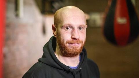 Billy Ramsay looks into the camera while standing in a boxing club - a punch bag can be seen blurred in the background. Billy has a bald head, a ginger beard and moustache and is wearing a black hoodie.