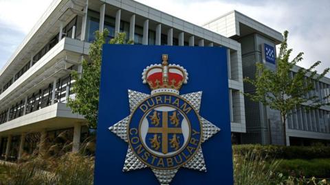 Durham Police's headquarters is a three-storey white building with tall windows. A big blue sign with Durham Constabulary's crest is in front of the building. 