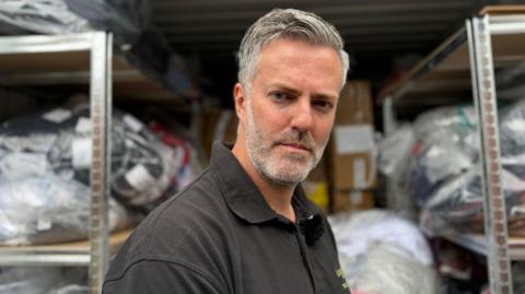 Dan Brown is standing in front of a container filled with bags of fake items seized by Lincolnshire Trading Standards. Dan is wearing a black T-shirt and has grey hair and a grey beard.