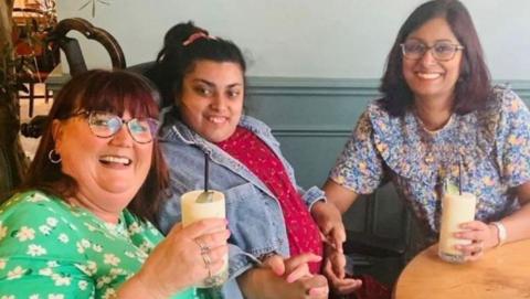 Three women sat around a table. Two with drinks in their hands either side of a woman in a wheelchair. One adult is wearing a green floral dress and has dark red hair and glasses, while another is wearing a blue floral dress with similar colour hair and also wearing glasses. The woman in the wheelchair is disabled and holding hands with the two other women. She has dark hair, a jean jacket and red top.