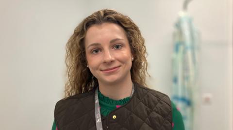 Olivia Howe looking at the camera, standing in a pale coloured room with a hospital gown hanging behind her