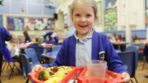 A primary school pupil in a light-blue polo shirt and royal blue cardigan is open-mouthed smiling at the camera while holding a red school dinner tray with spaghetti, broccoli and some sort of meat. There is also a section for cutlery, and a plastic beaker is obscuring her dessert. She has light blonde hair, pulled behind her head, in some sort of ponytail or bun (not seen in shot) and and has tendrils framing her face