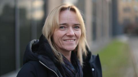 A woman with shoulder-length blonde hair smiles at the camera. She is wearing a black jacket and appears to be standing outside a building.