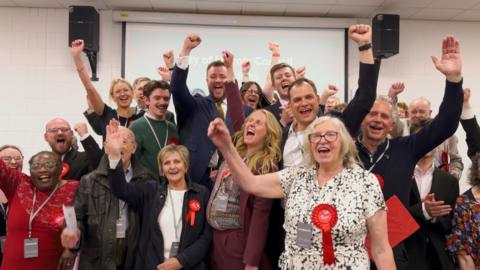 A group of people dressed in office attire and red badges cheering with their arms in the air. They are stood in a room with a powerpoint on display behind them. 