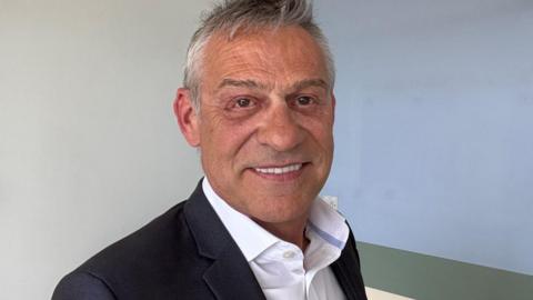 A man with grey hair is standing in front of a light coloured wall with a horizontal strip in a slightly darker shade. He is wearing a dark blazer over a white collared shirt.