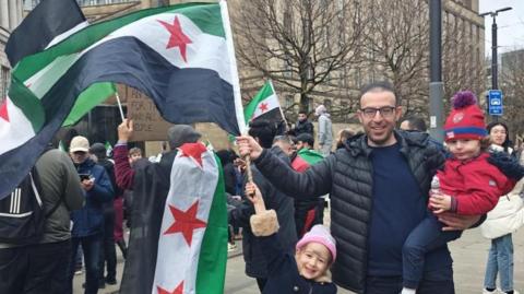 A man stand waving a Syrian flag on a street. He is holding a young boy in one arm and a young girl is standing on his other side also holding the bottom of the flag's handle. 