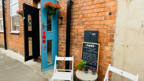 The outside entrance to nook gallery. There is a blue door wide open with the word nook painted in red. There is a small white table with two white deck chairs on either side.