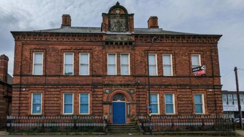 A two-storey red brick building with a central blue door and decorative stonework. The building has a “sold” sign displayed in an upper window.