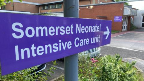 Close up of purple sign saying 'Somerset Neonatal Intensive Care Unit' in white lettering.  The entrance of the maternity ward and a car park are in the background.